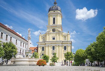 Radelnde am Karlsplatz in Neuburg an der Donau Radelnde auf dem Karlsplatz vor der prächtigen Hofkirche. Im Vordergrund ist ein Brunnen zu sehen.