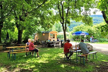 Mein Platz an der Bruck Blick auf den Schäferwagen und Rastplatz "Mein Platz an der Bruck". Im Vordergrund Bierbänke mit Gästen, im Hintergrund sieht man den umgebauten Schäferwagen und ankommende Radfahrer.