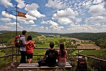 Urdonautalsteig bei Wellheim Vier Personen mit Rucksäcken blicken von einem Aussichtspunkt auf grüne Felder und bewaldete Hügel unter blauem Himmel.