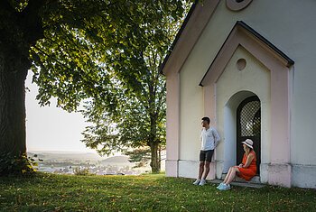 Wanderer auf der Monheimer Alb Mann lehnt an Hauswand, Frau sitzt in Türbogen, große Bäume, Dorf im Hintergrund bei Tageslicht.