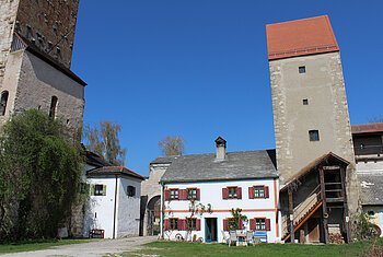 „Zu Gast im Denkmal“ in der Burg Nassenfels Bauernhof mit weißen Gebäuden, zwei Türmen und Gartenmöbeln unter blauem Himmel.