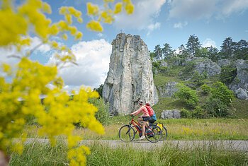 Radfahrer am Burgsteinfelsen Zwei Radfahrer fahren auf einem Weg vor einem hohen Felsen mit Kreuz, gelbe Blumen im Vordergrund, bewölkter Himmel