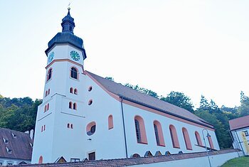 Wellheim Kath. Pfarrei St. Andreas Außenansicht gesamt Kirchturm mit Uhr an weißem Kirchengebäude vor bewaldetem Hügel unter hellem Himmel