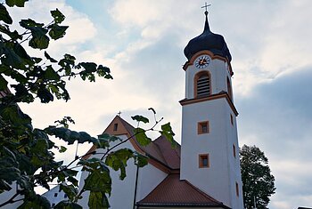 St. Johannes der Täufer Biesenhard bei Adelschlag Kirchturm mit Uhr und Kreuz, teilweise von Blättern vor bewölktem Himmel verdeckt.